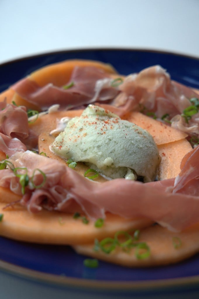 Entrée melon et jambon, sorbet vert et oignons nouveaux sur assiette bleue.