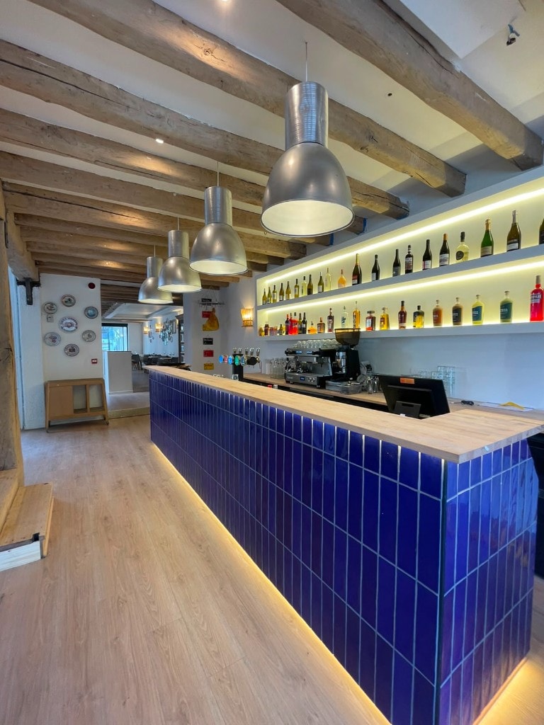Intérieur du bar de Jean & Gino à Reims, comptoir bleu, planches de bois et lumières industrielles sous une charpente en bois.